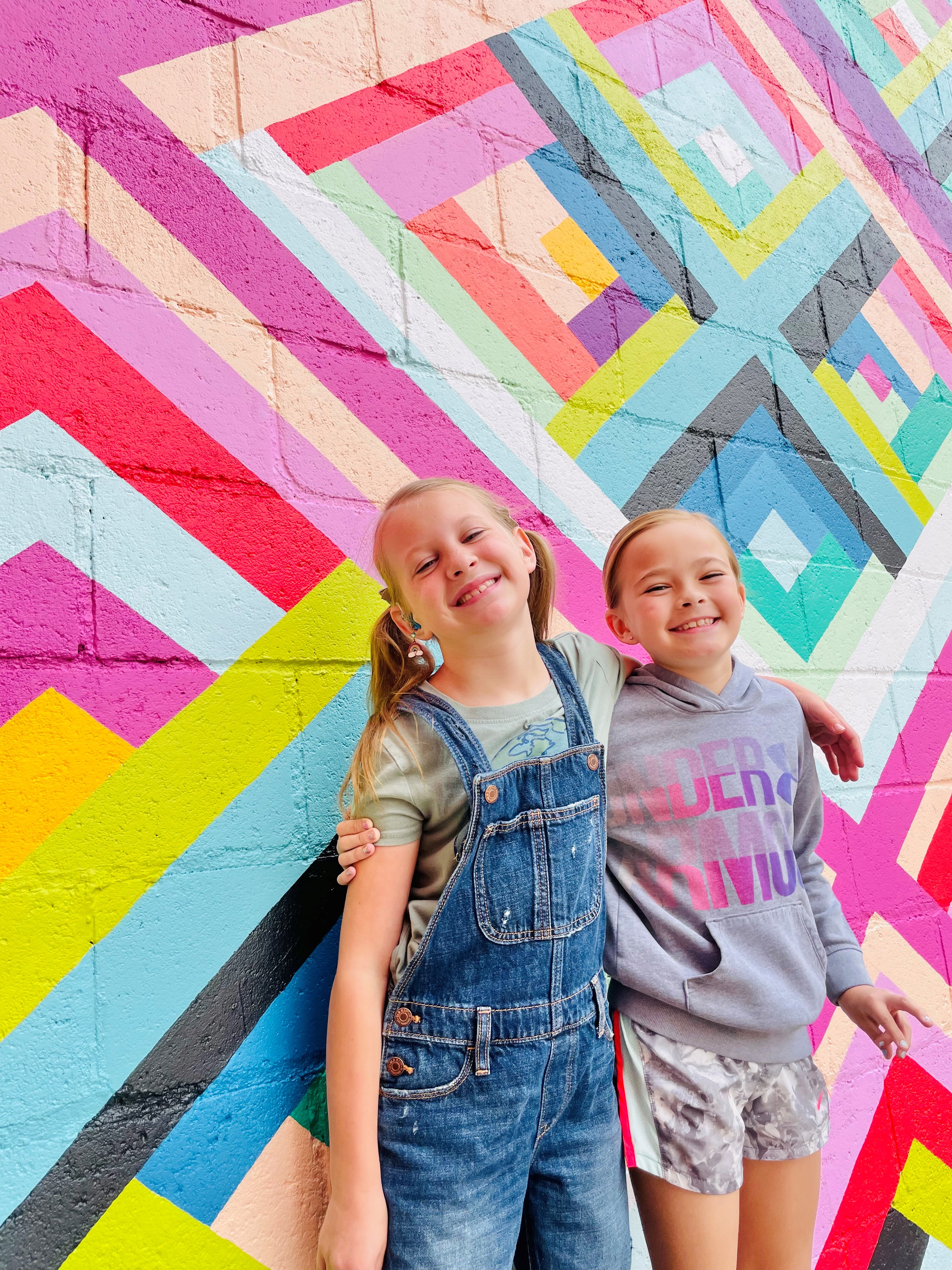 Two young girls hugging and smiling in front of a completed mural